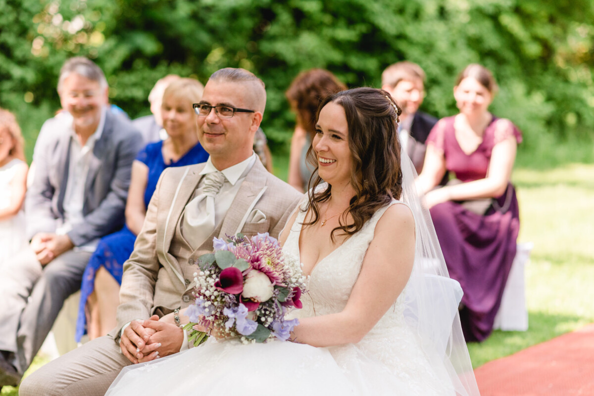 Braut und Bräutigam sitzen lächelnd zusammen, hinter ihnen sitzen Gäste im Freien. Die Braut hält einen Blumenstrauß.