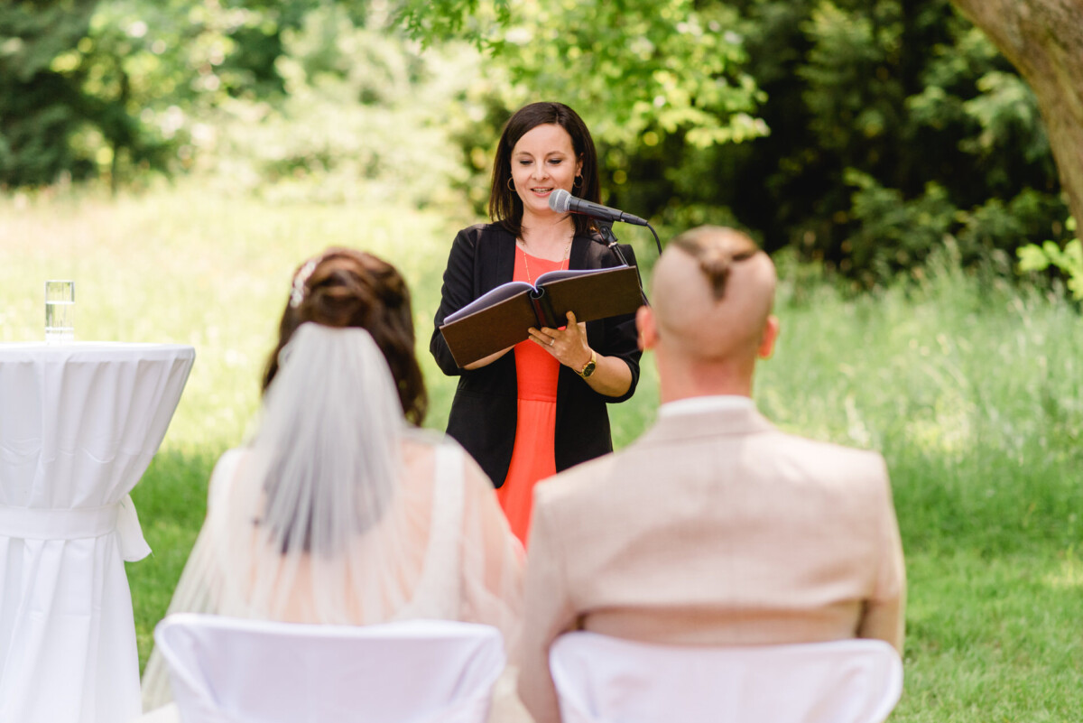 Eine Frau steht während einer Hochzeitszeremonie im Freien an einem Mikrofon und liest aus einem Buch vor. Vor ihr sitzen Braut und Bräutigam in einer grasbewachsenen Umgebung.