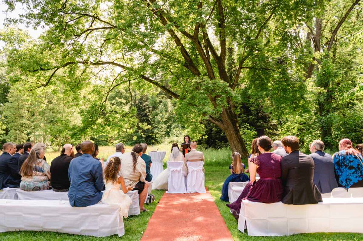 Hochzeitszeremonie im Freien mit Gästen, die an einem sonnigen Tag unter einem großen Baum auf weißen Bänken sitzen und einem Geistlichen gegenübersitzen.