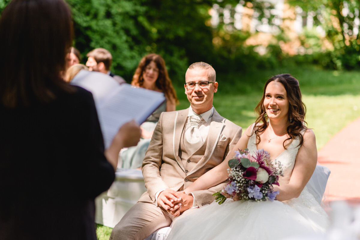 Ein Paar sitzt bei einer Hochzeitszeremonie im Freien nebeneinander. Die Braut hält einen Blumenstrauß und ein Geistlicher steht mit einem Buch vor ihnen.
