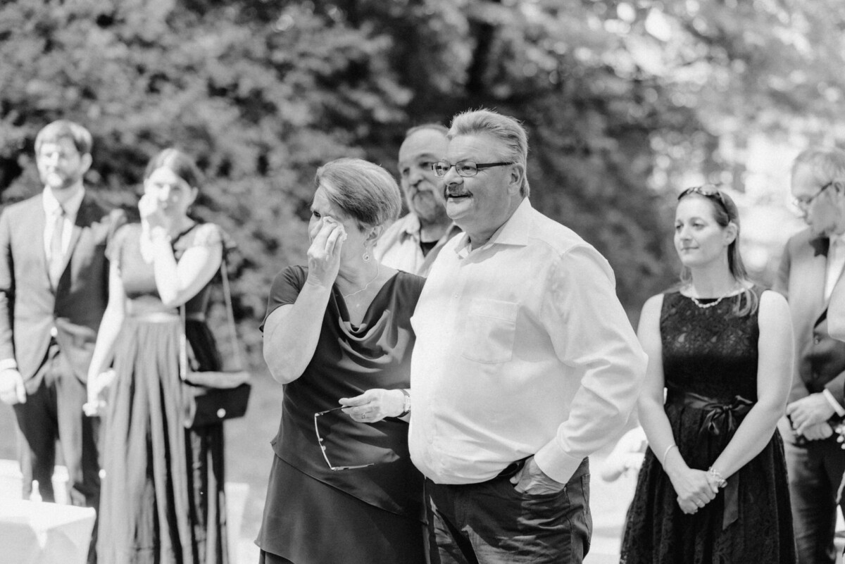 Eine Gruppe von Menschen steht im Freien bei einer Veranstaltung. Eine Frau wischt sich die Augen, während sie neben einem Mann steht. Andere im Hintergrund schauen zu.