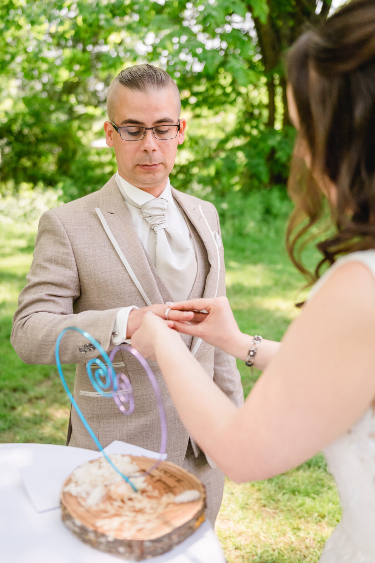 Ein Paar tauscht während einer Hochzeitszeremonie im Freien Ringe aus, im Hintergrund grüne Bäume.
