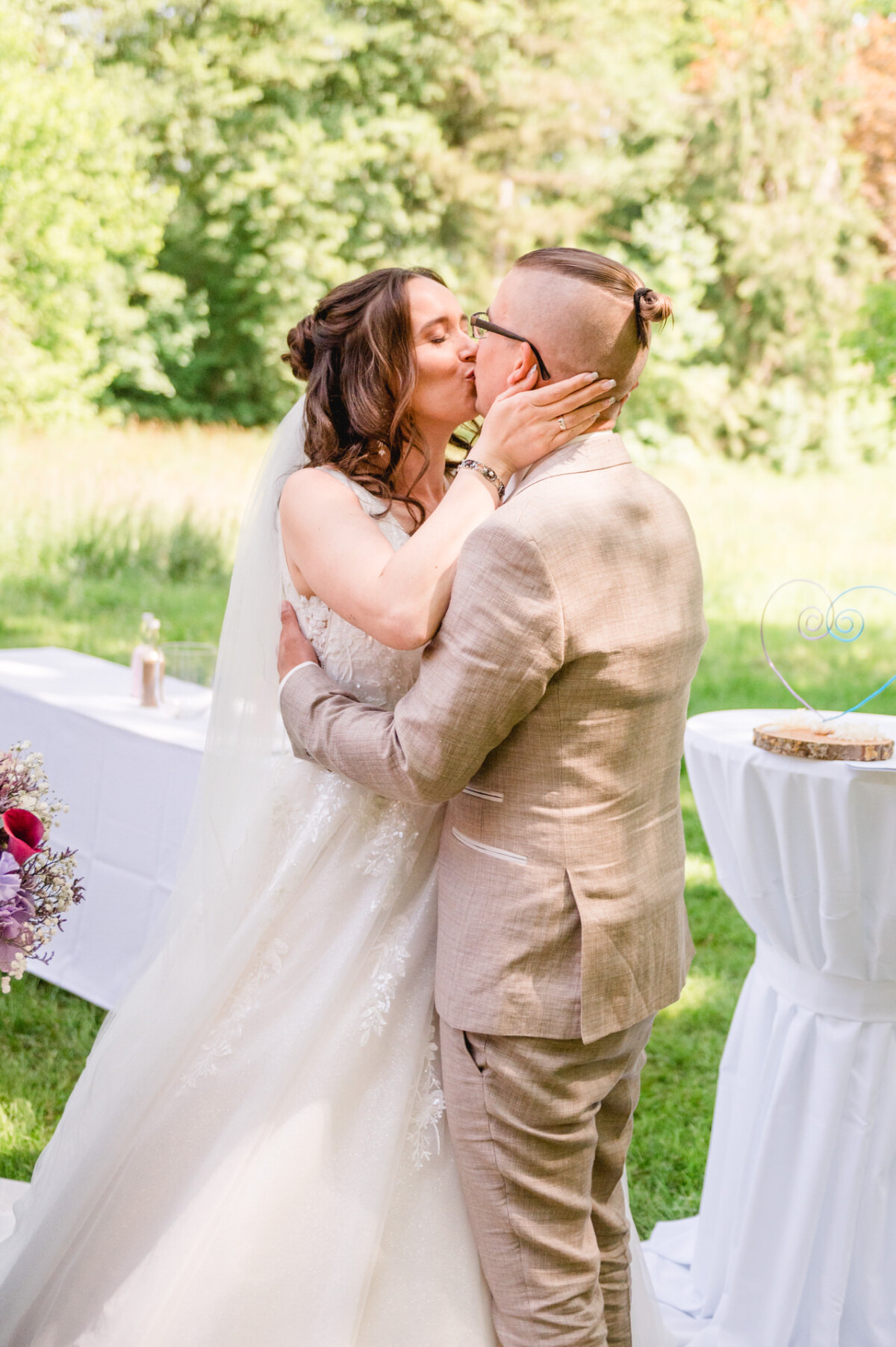 Braut und Bräutigam küssen sich während ihrer Hochzeitszeremonie im Freien, umgeben von Grün und dekorierten Tischen.