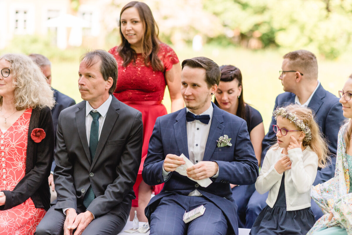 Eine Gruppe von Menschen, formell gekleidet, sitzt im Freien. Ein Mann in einem blauen Anzug hält ein Taschentuch und ein Mädchen mit Brille und Blumenkranz sitzt daneben und passt ihre Augen an.