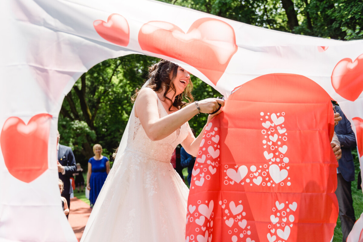 Während einer Hochzeitszeremonie im Freien schneidet eine Braut ein herzförmiges Muster aus einem Stoff mit Herzmotiven aus. Die Leute stehen in einer Gartenkulisse herum und beobachten die Szene.