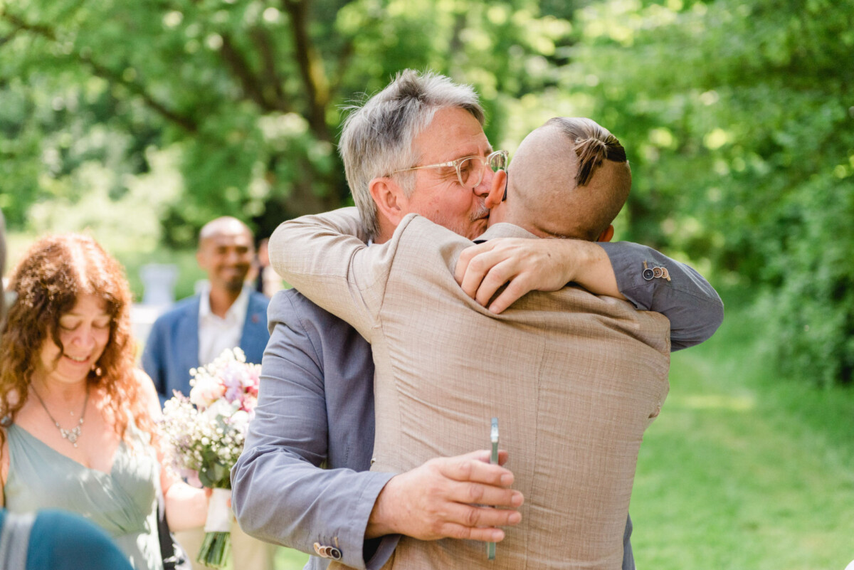 Zwei Männer umarmen sich im Freien, umgeben von Bäumen. Eine Frau mit Blumen steht lächelnd daneben.