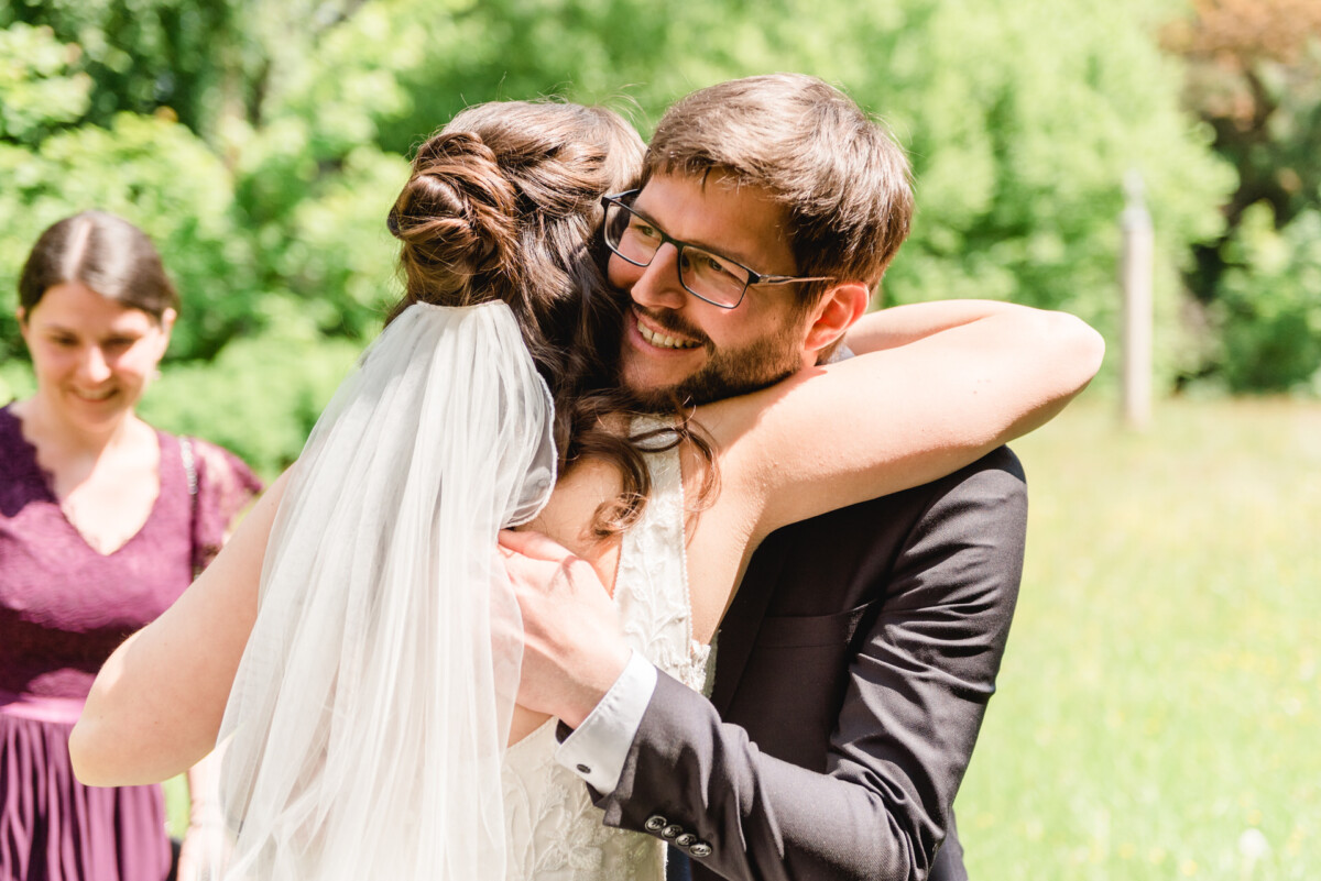 Eine Braut und ein Bräutigam umarmen sich an einem sonnigen Tag im Freien, im Hintergrund lächelt eine Frau in einem lila Kleid.