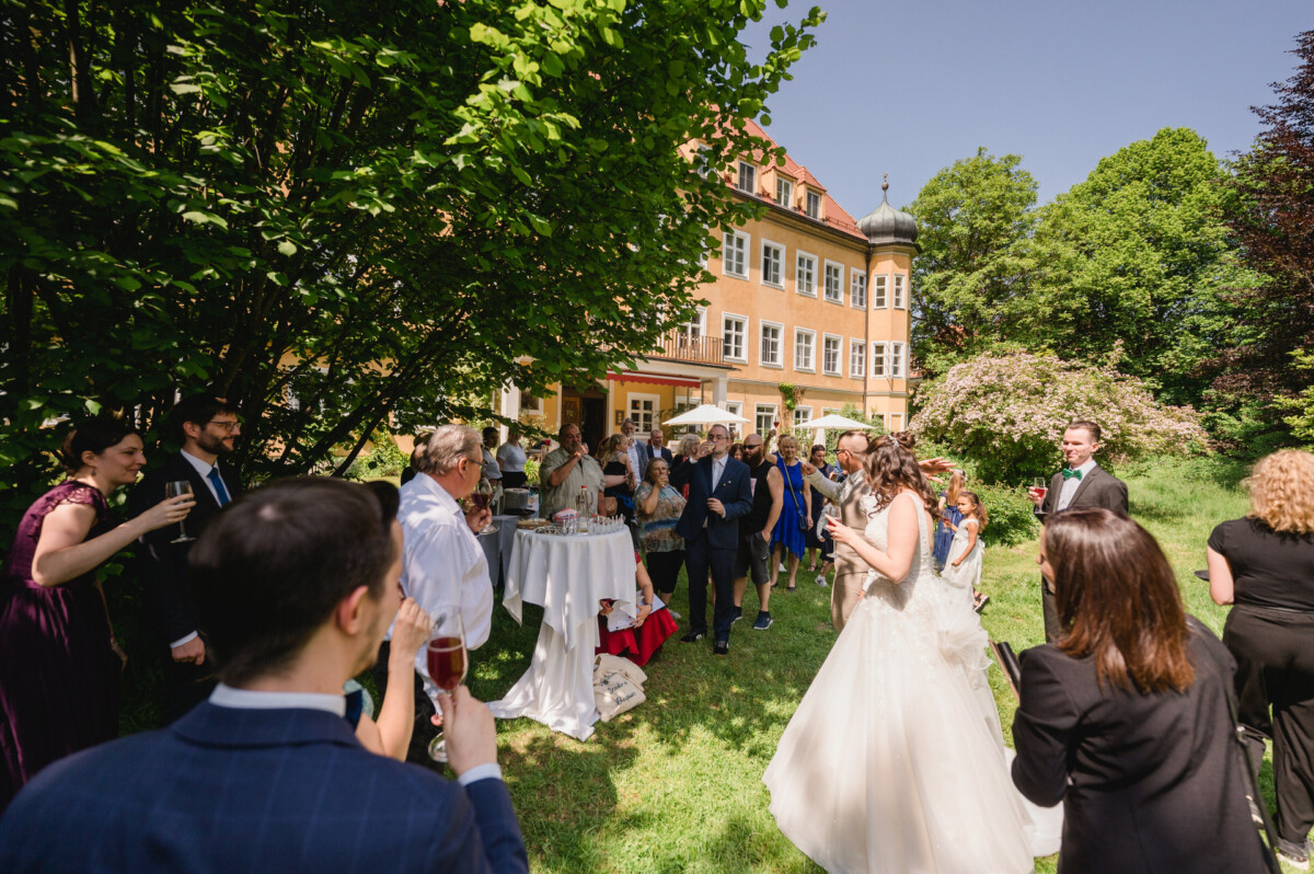 Menschen versammeln sich im Freien bei einer Hochzeitsfeier mit einem historischen Gebäude und Bäumen im Hintergrund. Gäste stehen, unterhalten sich und halten Getränke an runden Tischen.