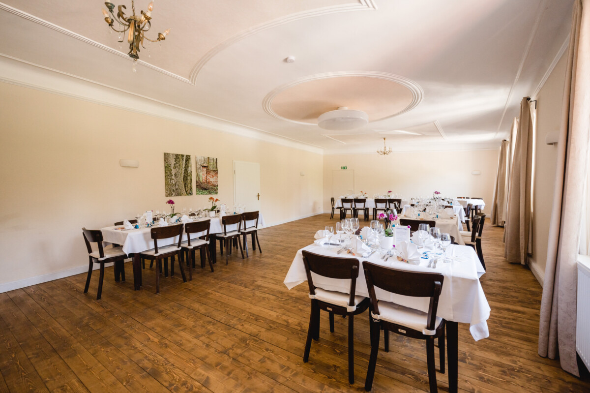 In einem hellen, leeren Esszimmer mit Holzfußboden sind mehrere Tische für ein formelles Essen mit weißen Tischdecken, Gläsern und Blumengestecken gedeckt.