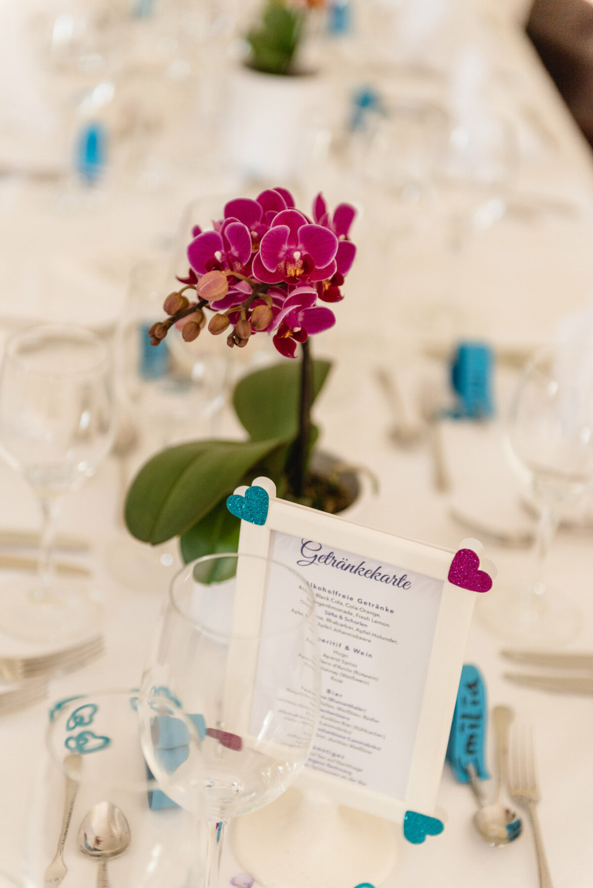 Ein Esstisch mit Glaswaren, Silberbesteck und einer violetten Orchidee als Tafelaufsatz. Im Vordergrund ist eine Speisekarte in einem Rahmen mit Herzdekorationen zu sehen.