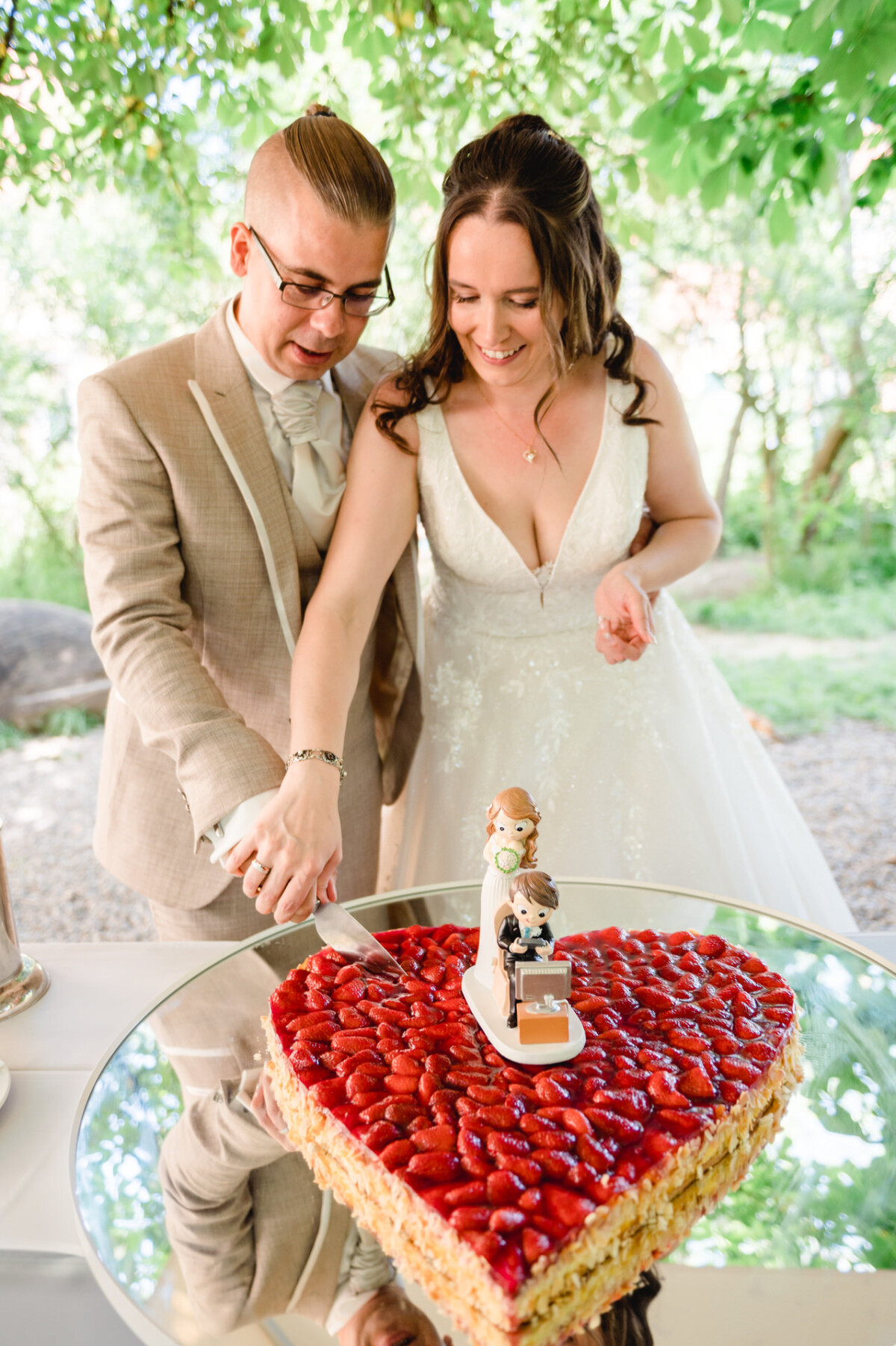 Ein Paar im Hochzeitskleid schneidet eine herzförmige Torte an, die mit Erdbeeren und einer kleinen Braut- und Bräutigamfigur garniert ist.