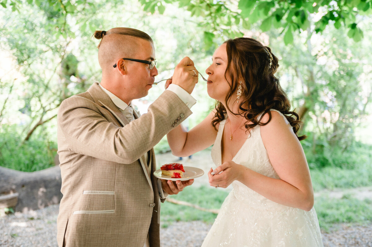 Ein Paar im Hochzeitskleid füttert sich gegenseitig im Freien unter einem Baum mit Kuchen.