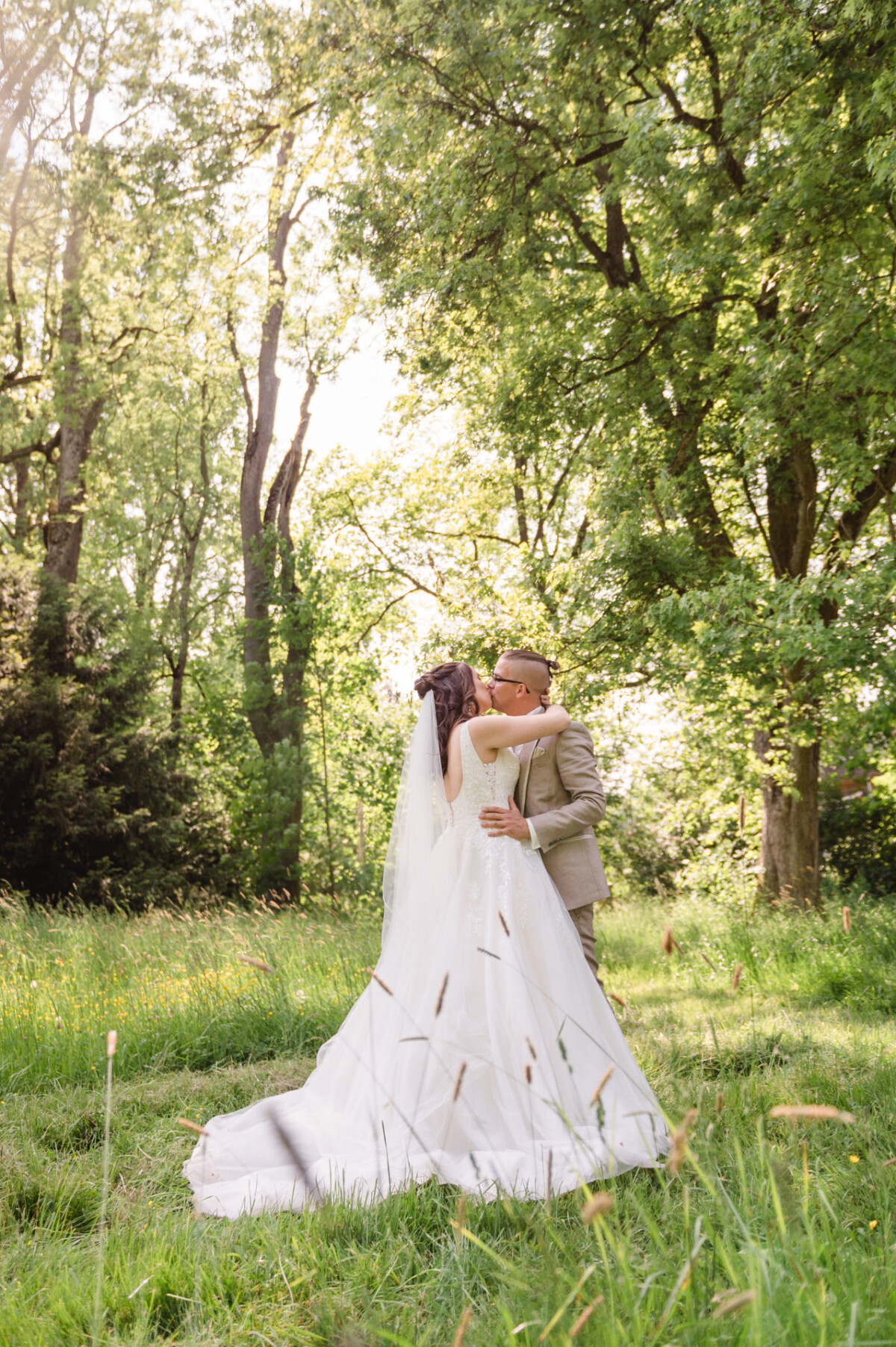 Ein Paar im Hochzeitskleid umarmt und küsst sich auf einer sonnenbeschienenen, von Bäumen umgebenen Wiese.