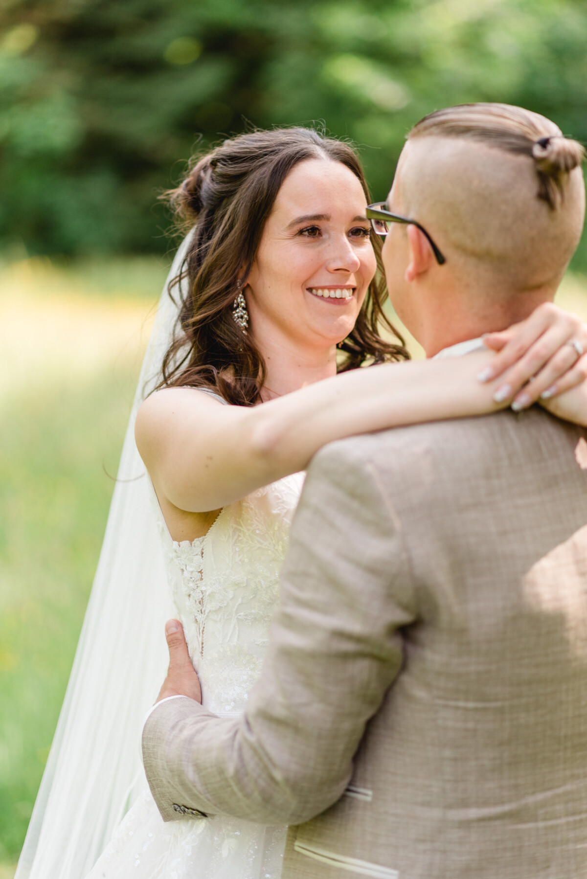 Ein Paar umarmt sich im Freien. Die Braut trägt ein weißes Kleid und einen Schleier und der Bräutigam einen hellbraunen Anzug vor einem grünen Hintergrund.
