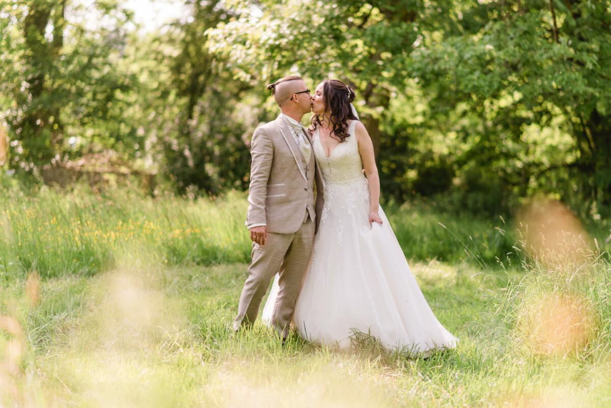 Ein Paar in Abendgarderobe küsst sich im Freien auf einer Rasenfläche mit Bäumen und Sonnenlicht im Hintergrund.