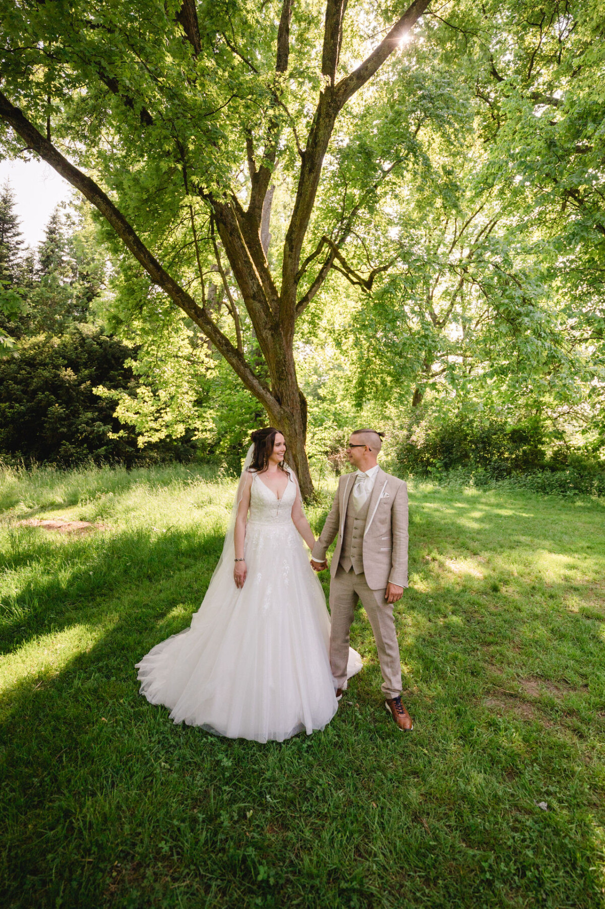 Ein Paar im Hochzeitskleid hält Händchen unter einem großen Baum mit üppigem grünem Laub.