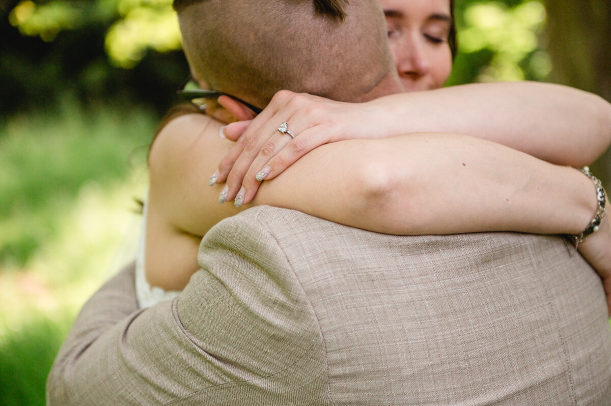 Zwei Menschen umarmen sich im Freien; einer trägt einen beigen Anzug, der andere zeigt einen Verlobungsring.