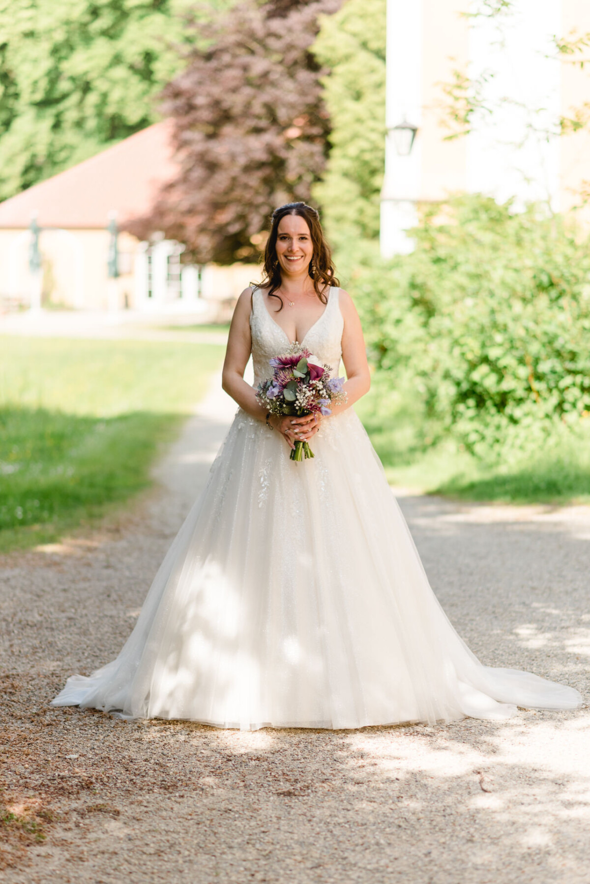 Eine Braut im weißen Kleid hält einen Blumenstrauß in der Hand, während sie auf einem Kiesweg in einer Gartenanlage steht.