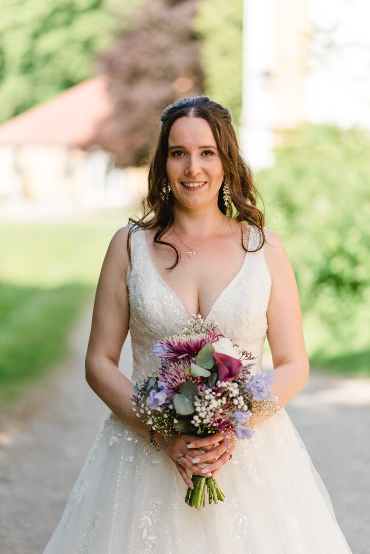 Eine Braut in einem weißen Kleid und einem bunten Blumenstrauß steht draußen auf einem Weg, im Hintergrund ist Grün zu sehen.