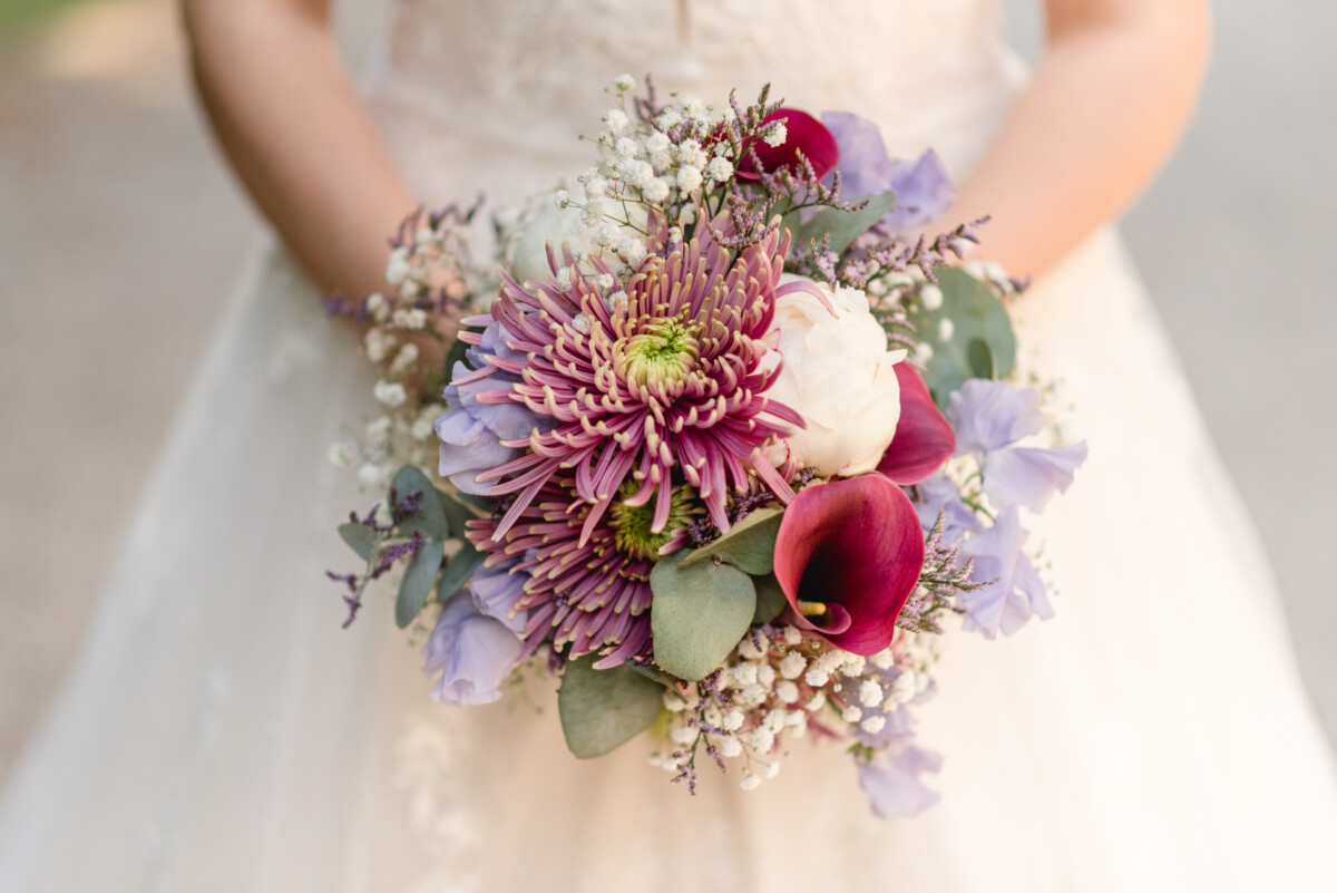 Eine Person hält einen Blumenstrauß mit violetten Astern, weißen Pfingstrosen, roten Callas, Schleierkraut und Grünpflanzen vor einem unscharfen Hintergrund.