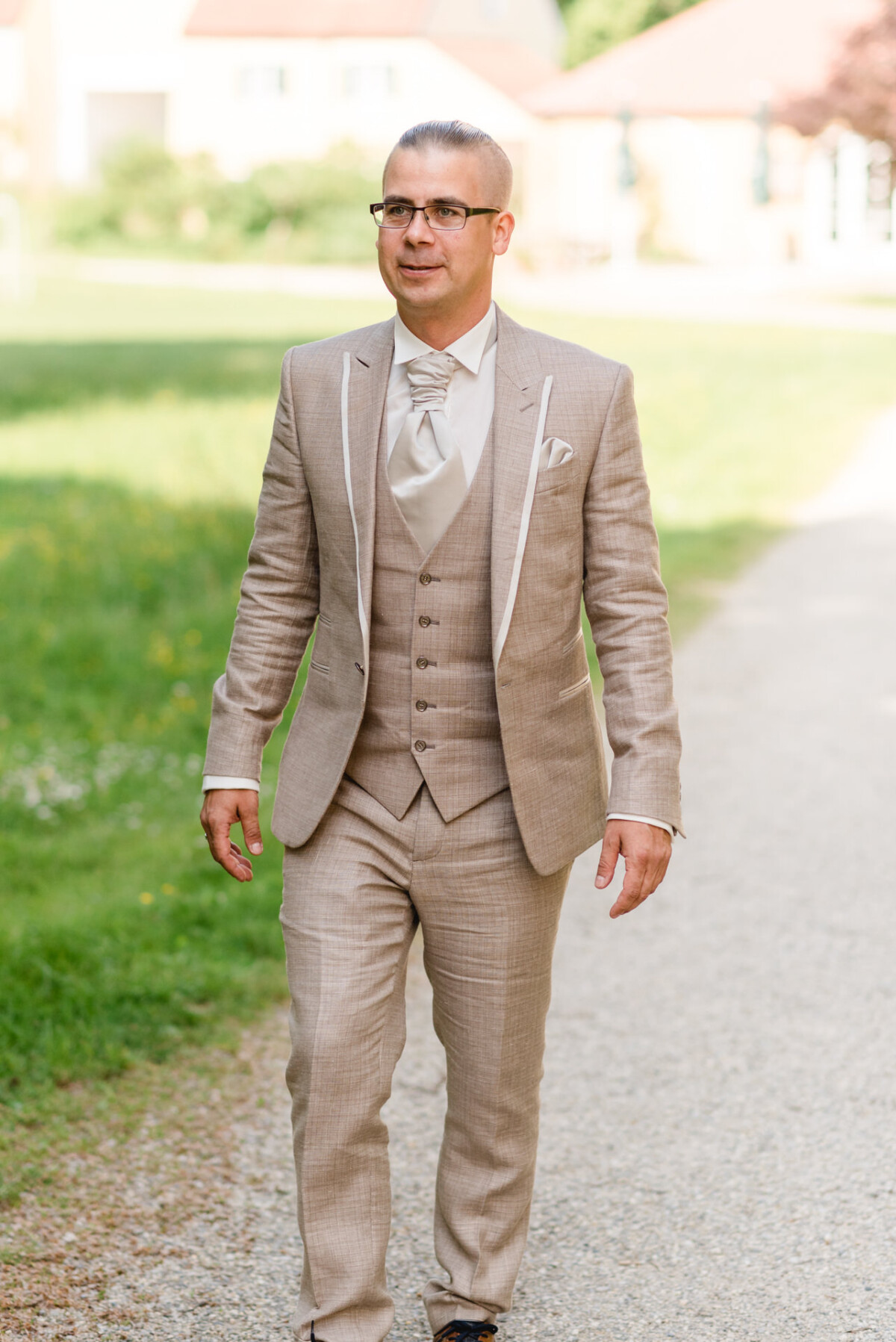 Ein Mann in einem hellbraunen Anzug geht im Freien auf einem Kiesweg.