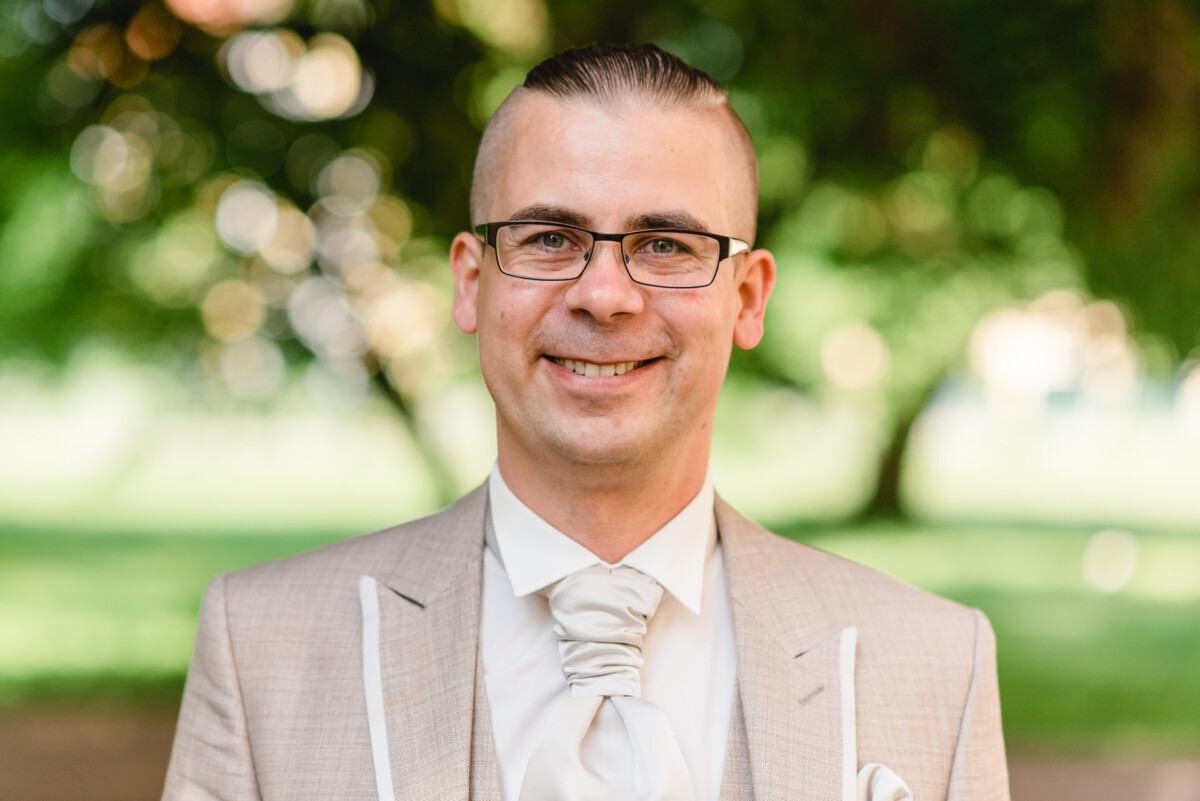 Ein Mann mit Brille, beigem Anzug und weißer Krawatte lächelt im Freien mit verschwommenem Grün im Hintergrund.