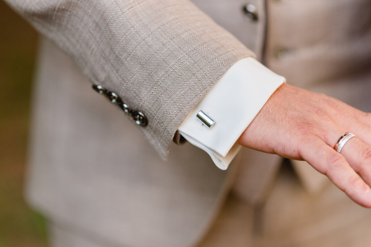 Nahaufnahme einer Person, die einen beigen Anzug mit weißem Hemd trägt. An der Hemdmanschette befindet sich ein silberner Manschettenknopf und an der Hand ein Ring.