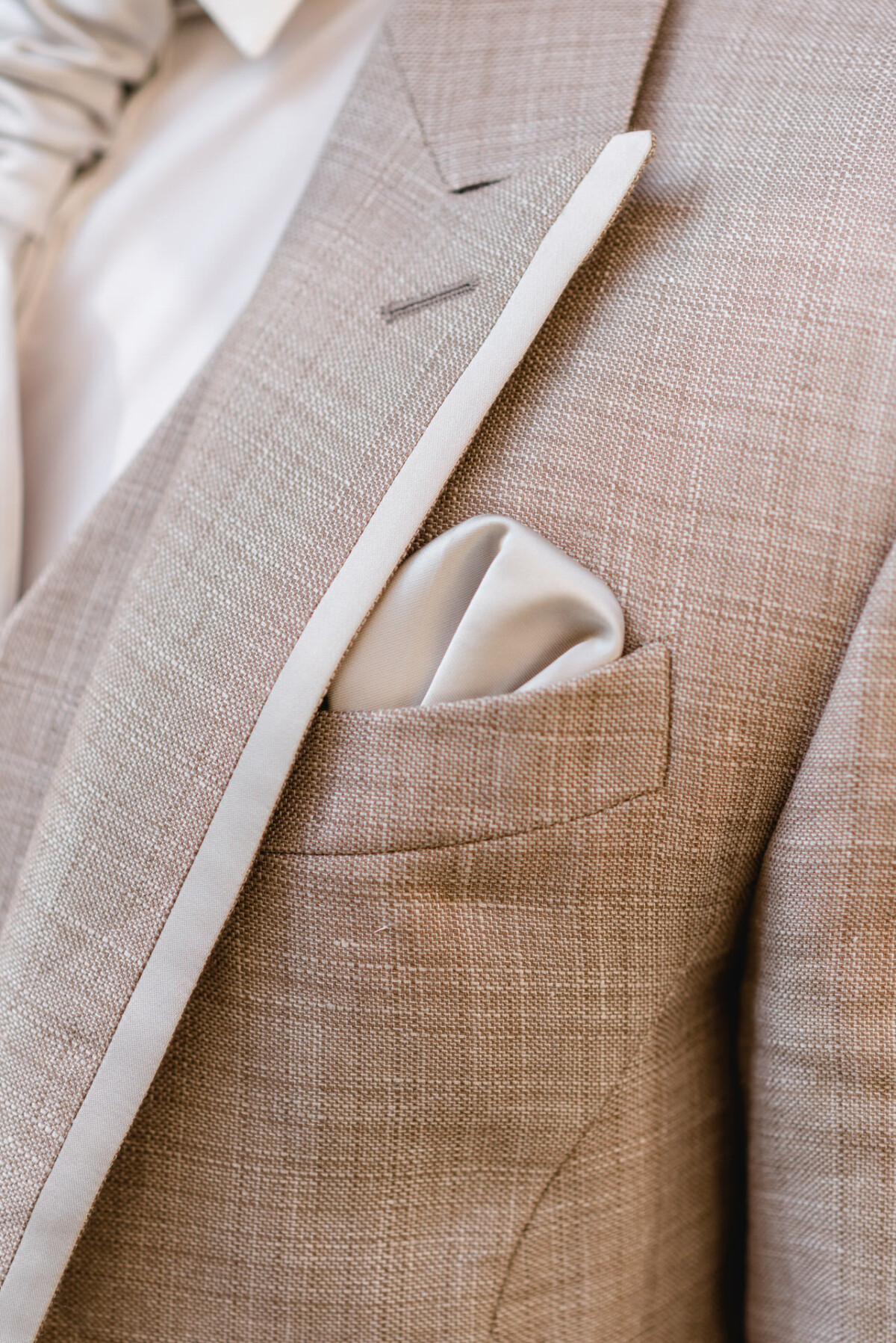 Nahaufnahme eines beigen Anzugs mit einem weißen Einstecktuch in der Brusttasche und einem darunter sichtbaren weißen Hemd.