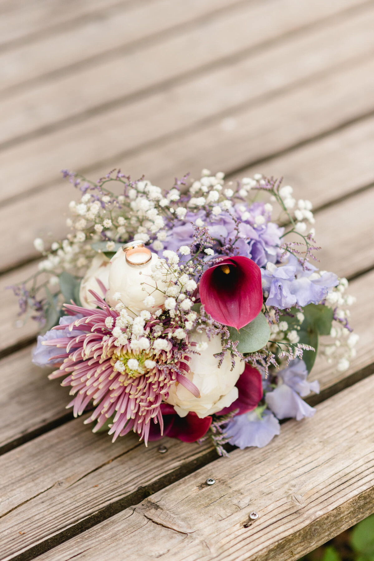 Ein Strauß mit weißen Pfingstrosen, lila Callas, rosa Chrysanthemen, Schleierkraut und Grünpflanzen auf einer Holzoberfläche.