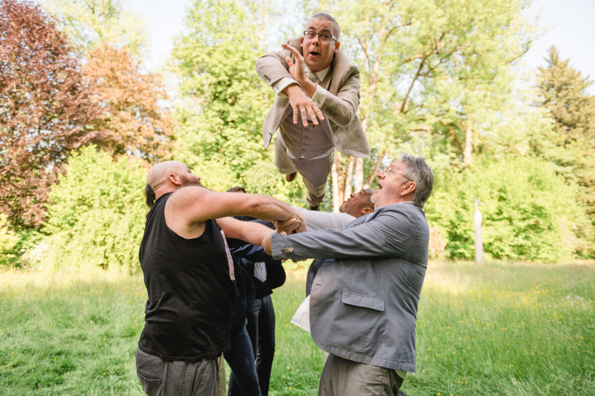Drei Männer werfen einen anderen Mann in einem beigen Anzug in einer grasbewachsenen Parklandschaft in die Luft.
