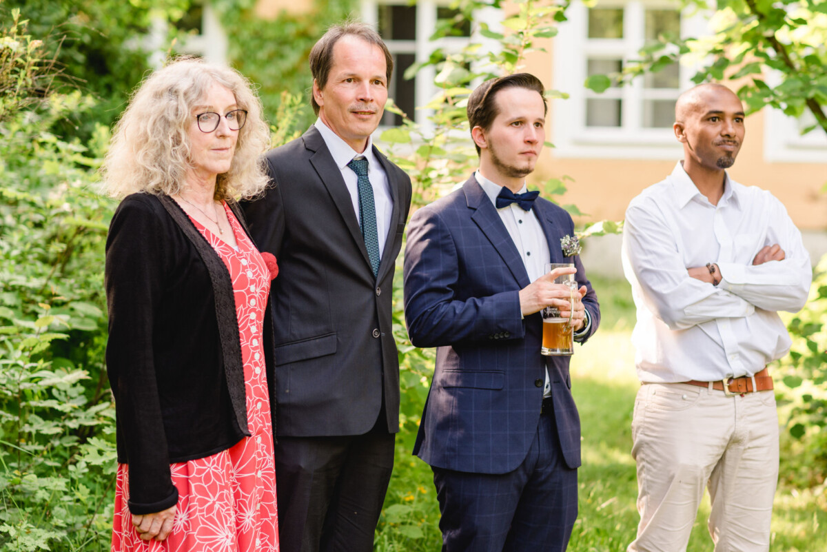 Vier Personen stehen im Freien und blicken in die gleiche Richtung. Einer hält ein Glas Bier. Sie sind in formelle Kleidung gekleidet und von Grün umgeben.