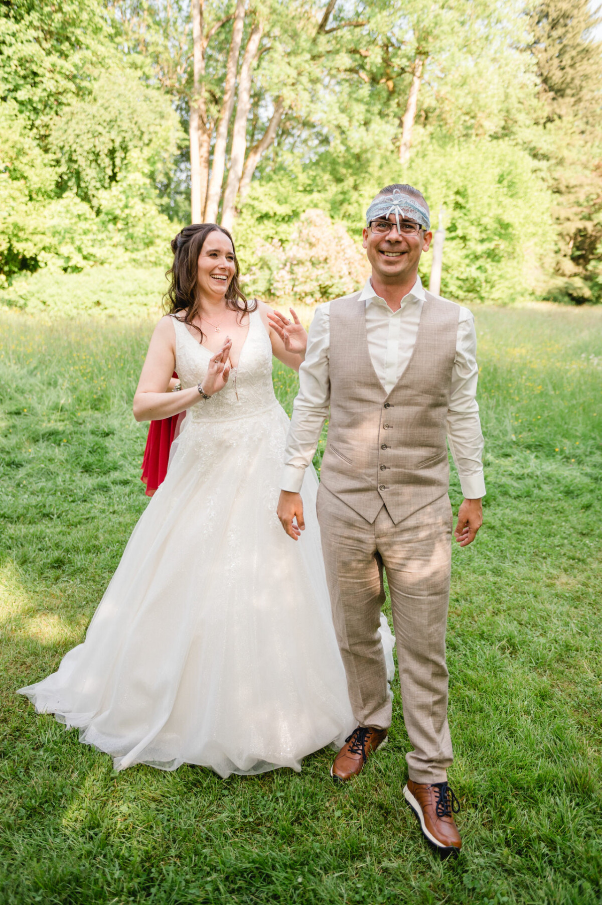 Eine Braut überrascht ihren Bräutigam mit verbundenen Augen spielerisch in einer grasbewachsenen, sonnenbeschienenen Umgebung im Freien.