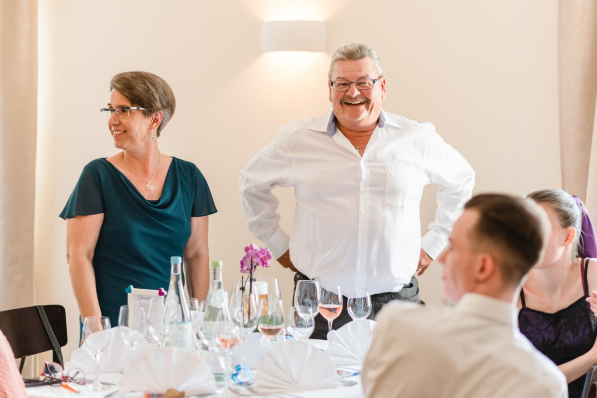 Ein Mann und eine Frau stehen lächelnd neben einem Esstisch mit Weingläsern und dekorativen Servietten.