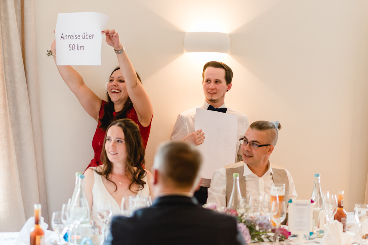 Menschen sitzen an einem Esstisch, eine Frau steht da und hält ein Schild mit der Aufschrift „Anreise über 50 km“.