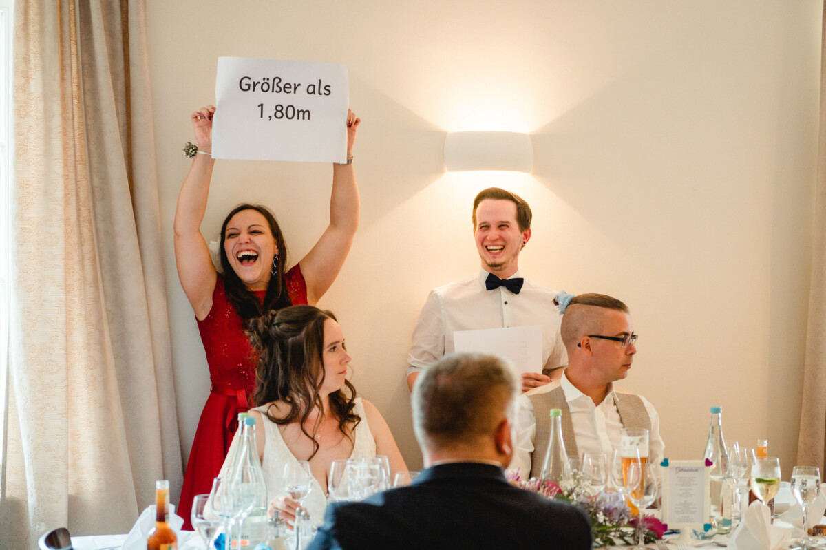 Zwei Menschen stehen lächelnd an einem Esstisch. Einer hält ein Schild mit der Aufschrift „Größer als 1,80m“. Drei sitzende Menschen sind zu sehen, umgeben von Flaschen und Gläsern.