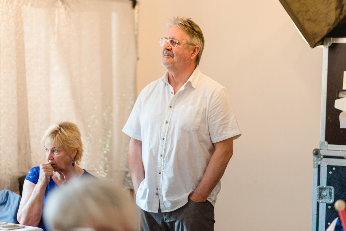 Ein Mann im weißen Hemd steht mit den Händen in den Taschen neben einer sitzenden Frau in Blau in einem Raum.