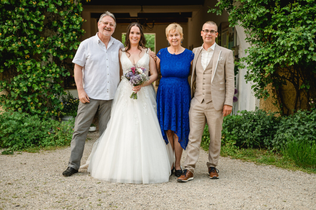 Vier Personen stehen zusammen vor einem grünen Strauch und einer Tür. Eine Braut in einem weißen Kleid hält Blumen, flankiert von zwei Männern und einer Frau, alle in Abendgarderobe gekleidet.