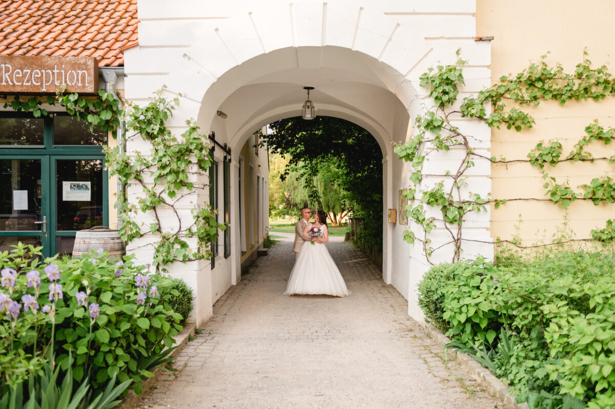 Eine Braut in einem weißen Kleid steht mit einem Blumenstrauß in der Hand unter einem weinbewachsenen Torbogen an einem Veranstaltungsort im Freien.
