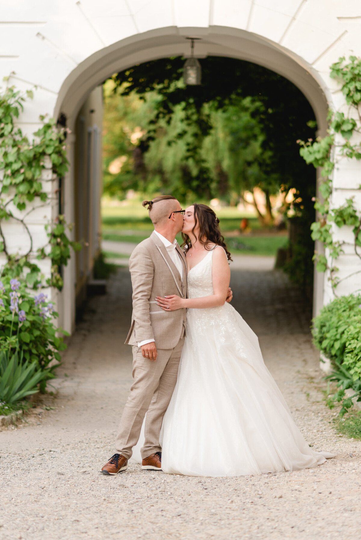 Braut und Bräutigam küssen sich unter einem mit Grün geschmückten Torbogen. Die Braut trägt ein weißes Kleid, der Bräutigam einen hellbraunen Anzug. Im Hintergrund sind ein Weg und ein Garten zu sehen.