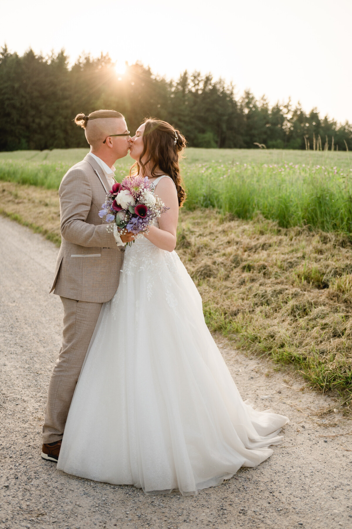 Ein Paar küsst sich auf einer unbefestigten Straße, umgeben von Grün. Die Person links trägt einen hellbraunen Anzug und die Person rechts ein weißes Hochzeitskleid und hält einen Blumenstrauß.