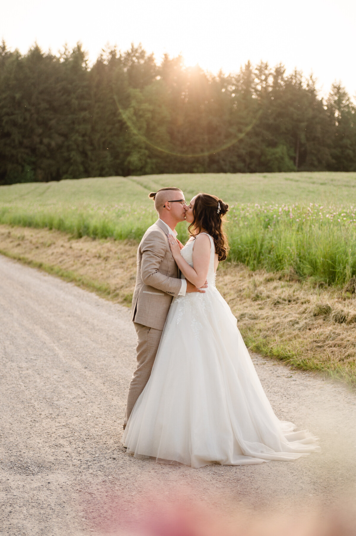 Ein Paar in Hochzeitskleidung steht auf einem Kiesweg, umgeben von Grün, unter einem klaren Himmel, während die Sonne hinter den Bäumen untergeht. Sie umarmen sich und schauen sich an.