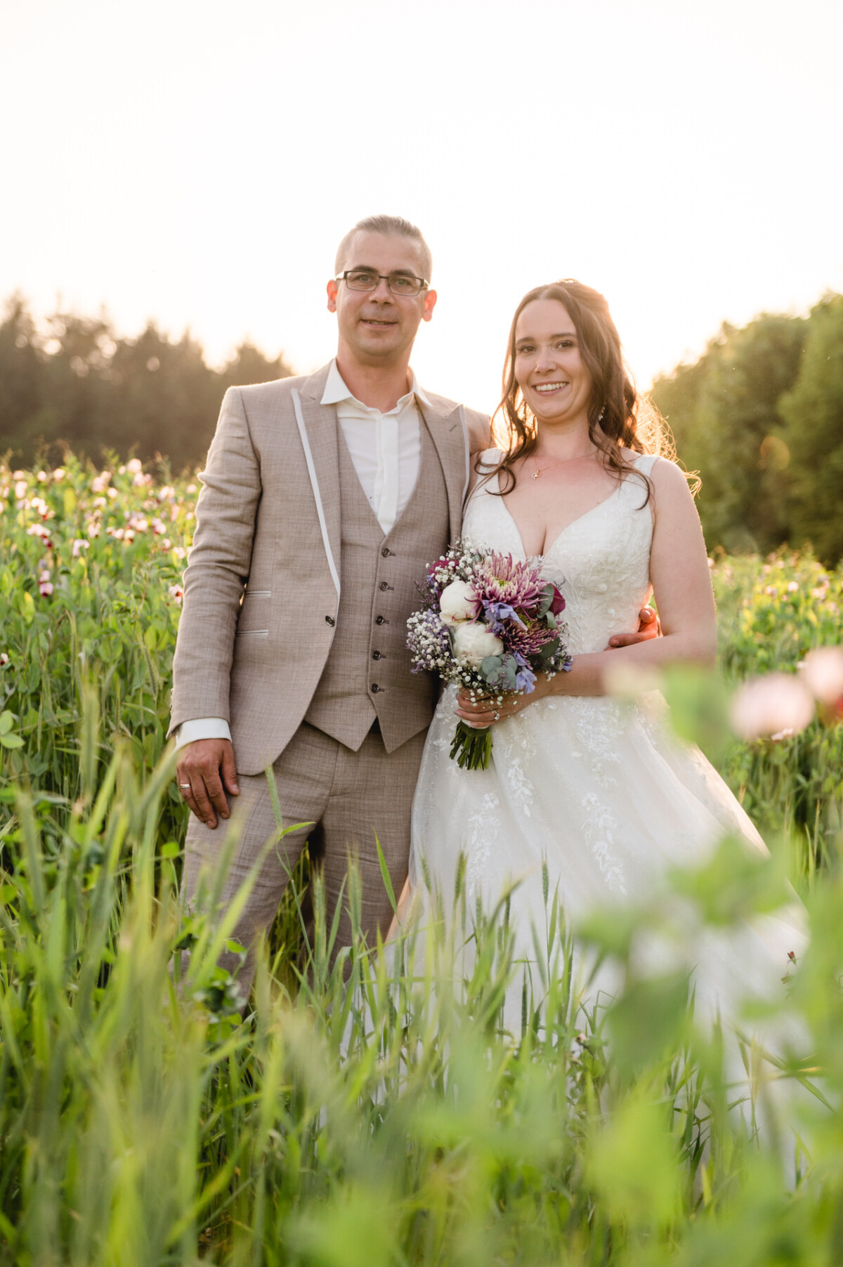 Ein Paar steht auf einem Feld mit hohem Gras. Der Mann trägt einen hellbraunen Anzug und die Frau in einem weißen Brautkleid hält einen Blumenstrauß in der Hand. Im Hintergrund geht die Sonne unter.