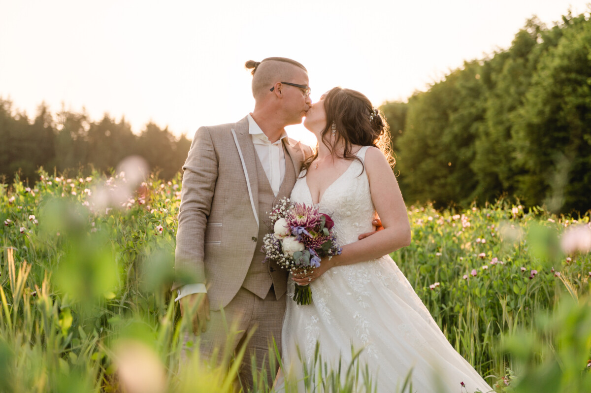Ein Paar im Hochzeitskleid steht bei Sonnenuntergang auf einem Feld und küsst sich. Die Braut hält einen Blumenstrauß.