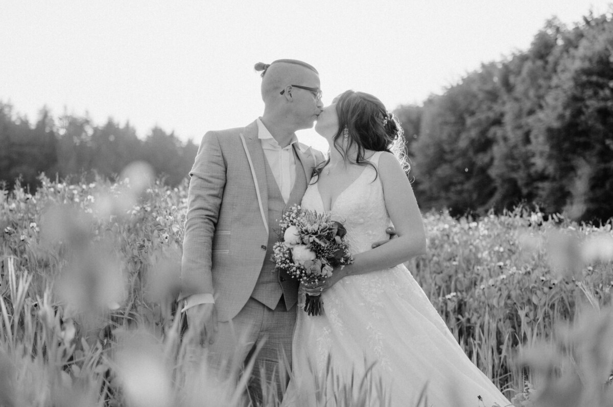Ein Paar in Hochzeitskleidung steht auf einem Feld mit hohem Gras, während der Bräutigam die Stirn der Braut küsst. Die Braut hält einen Blumenstrauß und im Hintergrund sind Bäume zu sehen. Das Foto ist schwarzweiß.