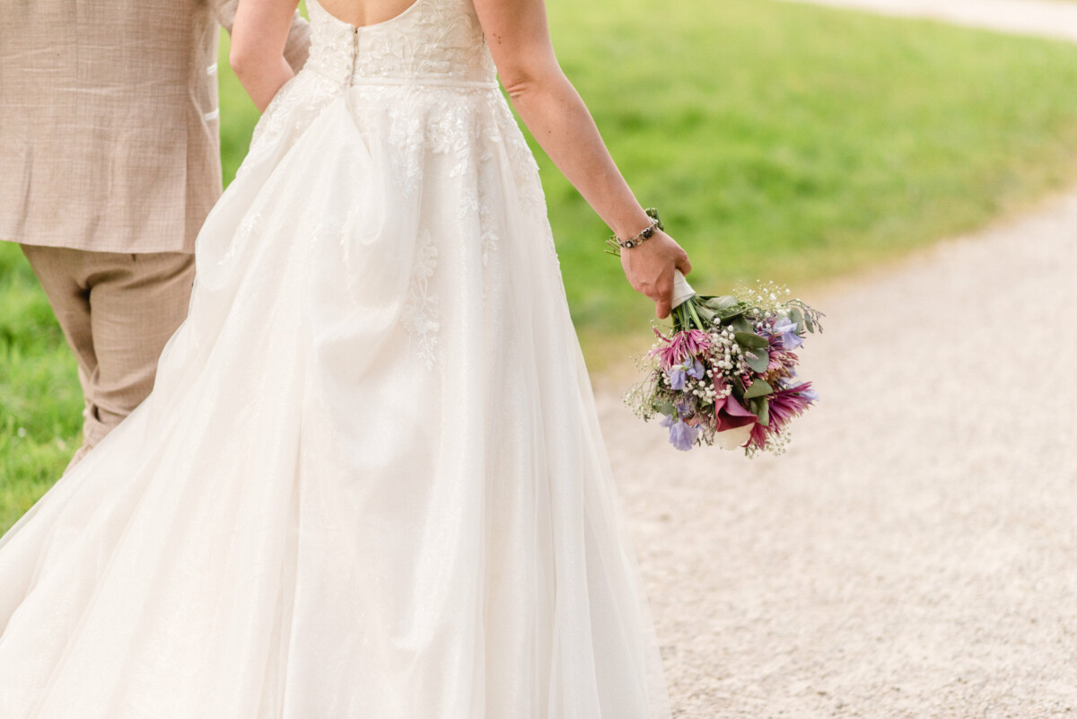 Eine Braut im weißen Kleid hält einen Blumenstrauß, während sie mit einem Bräutigam im Anzug auf einem Kiesweg spazieren geht.