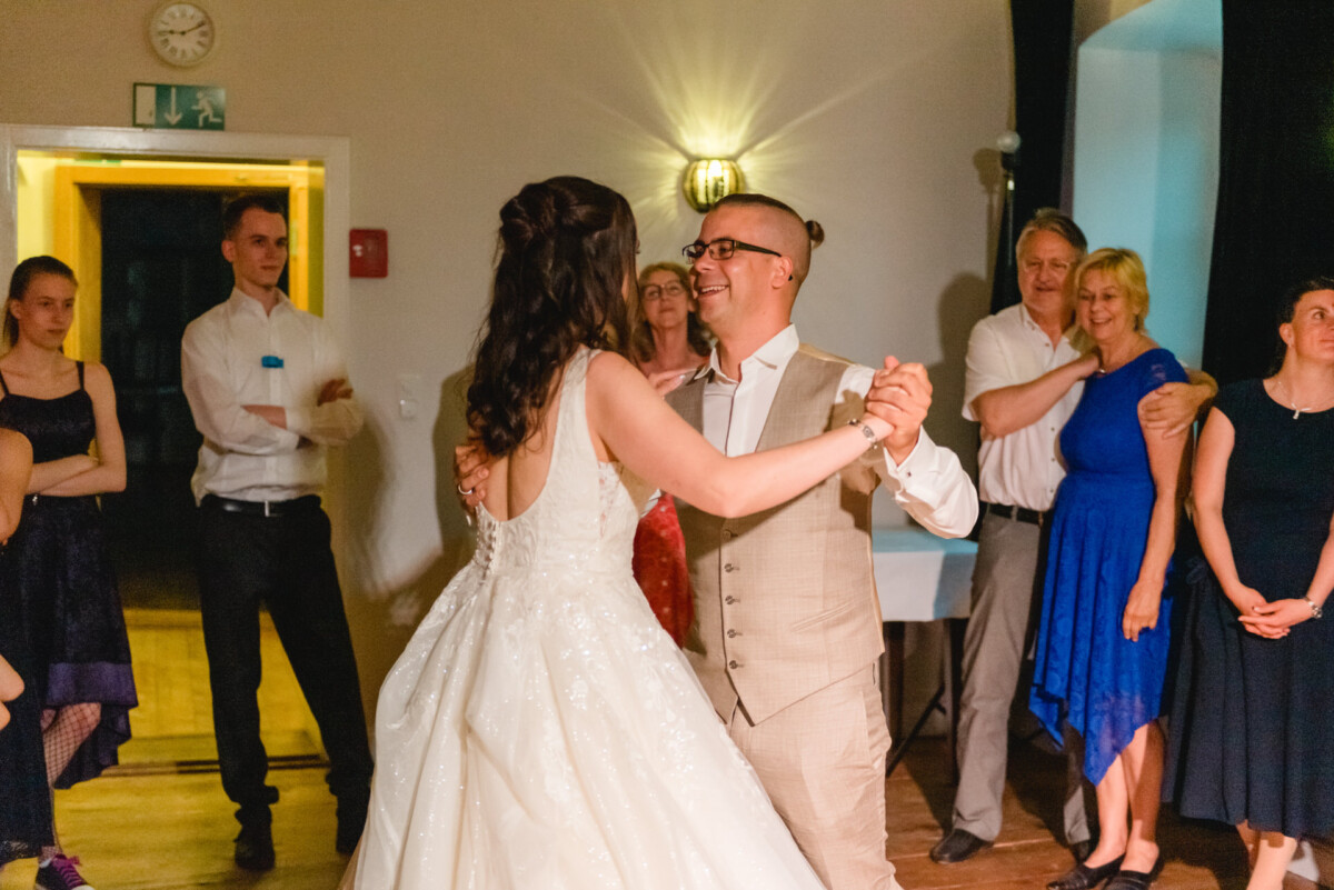 Ein Paar tanzt bei einer Indoor-Veranstaltung. Der Mann trägt einen beigen Anzug und die Frau ein weißes Kleid. Mehrere Leute stehen herum und schauen zu.
