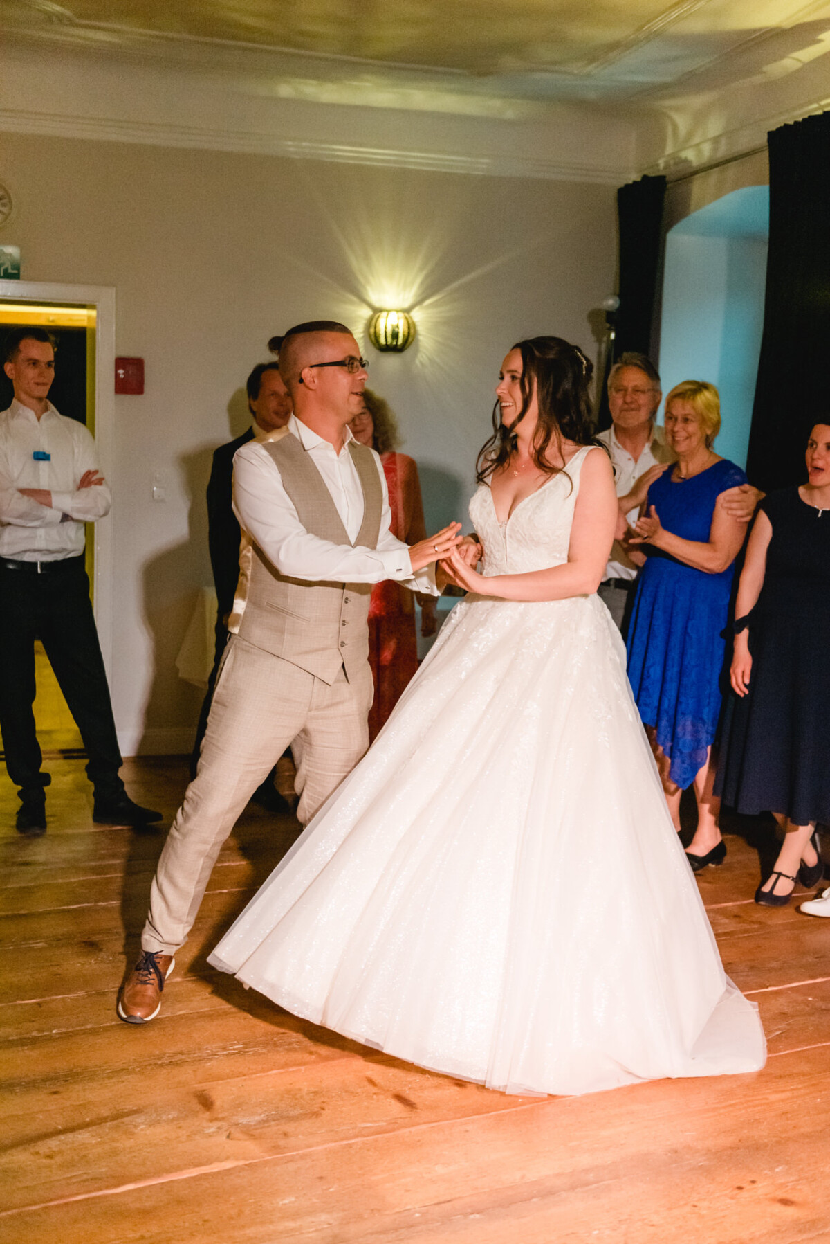 Ein Paar tanzt bei einer Hochzeitsfeier. Der Bräutigam trägt einen beigen Anzug und die Braut ein weißes Kleid. Die Gäste stehen auf einem Holzboden herum und schauen zu.
