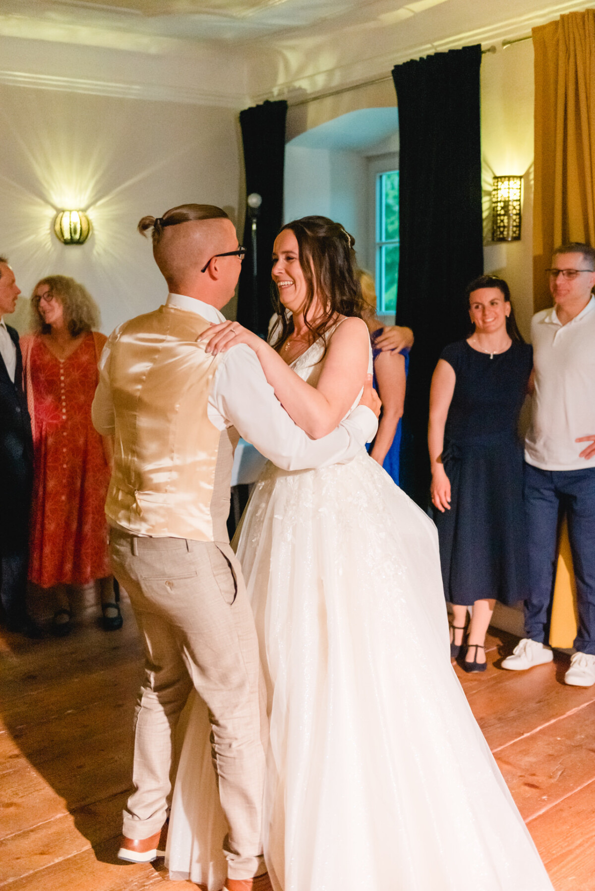 Braut und Bräutigam tanzen zusammen in einem warm beleuchteten Raum, umgeben von lächelnden Gästen.
