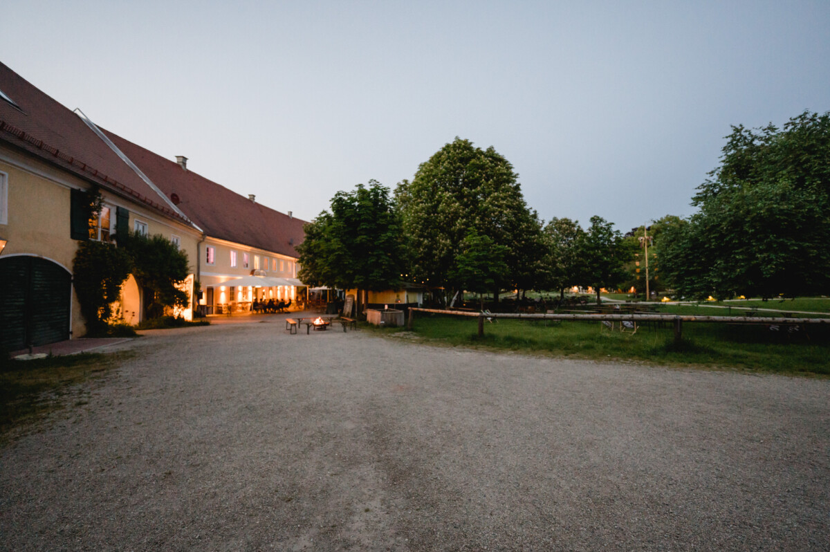 Großer Innenhof mit Kiesboden, links von einem langen Gebäude mit erleuchteten Fenstern begrenzt. Im Hintergrund sind Bäume und ein Zaun. Das Abendlicht sorgt für eine ruhige Atmosphäre.