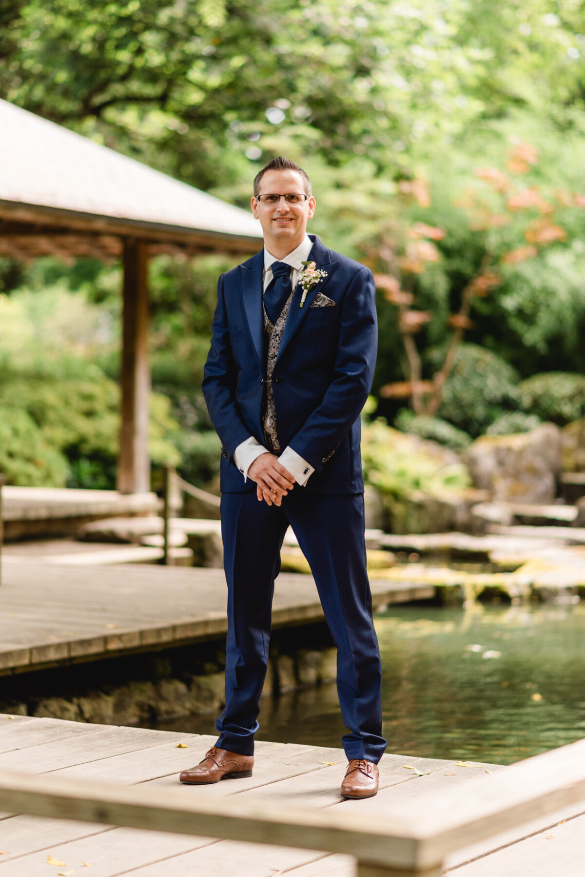 Ein Mann im blauen Anzug steht auf einer Holzbrücke in einer Gartenlandschaft, umgeben von Grün und einem kleinen Teich.
